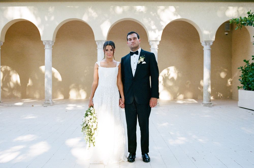 Bride and groom stand elegantly under arched colonnade, she in white gown with bouquet, he in black tuxedo with bow tie.