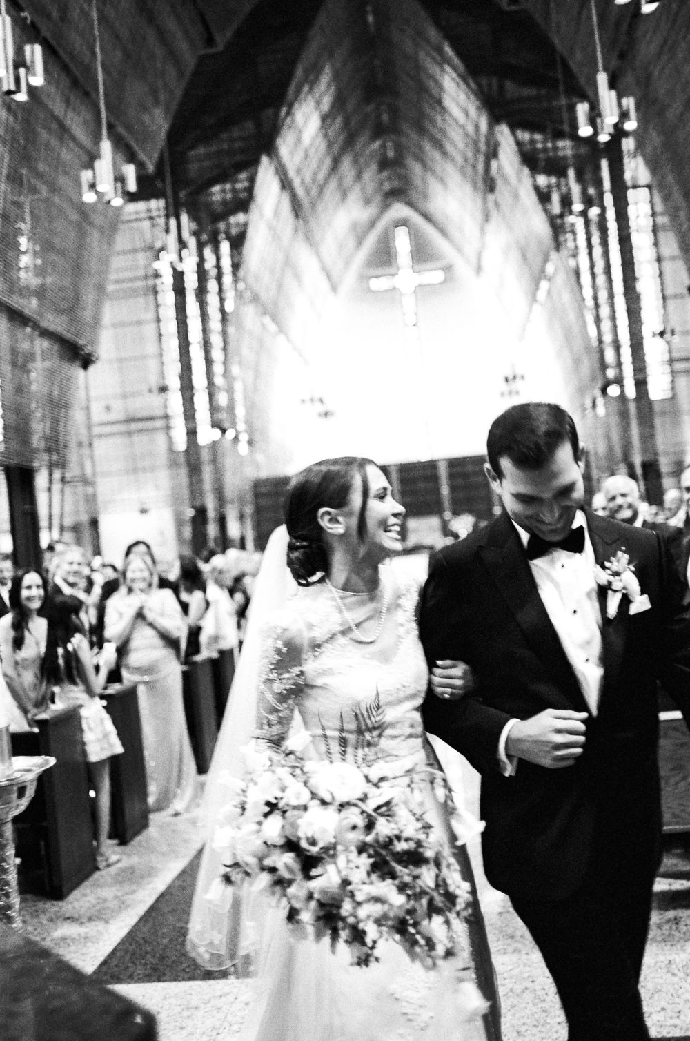 Black and white photo of a smiling bride and groom walking down the aisle of a grand church after their wedding ceremony.