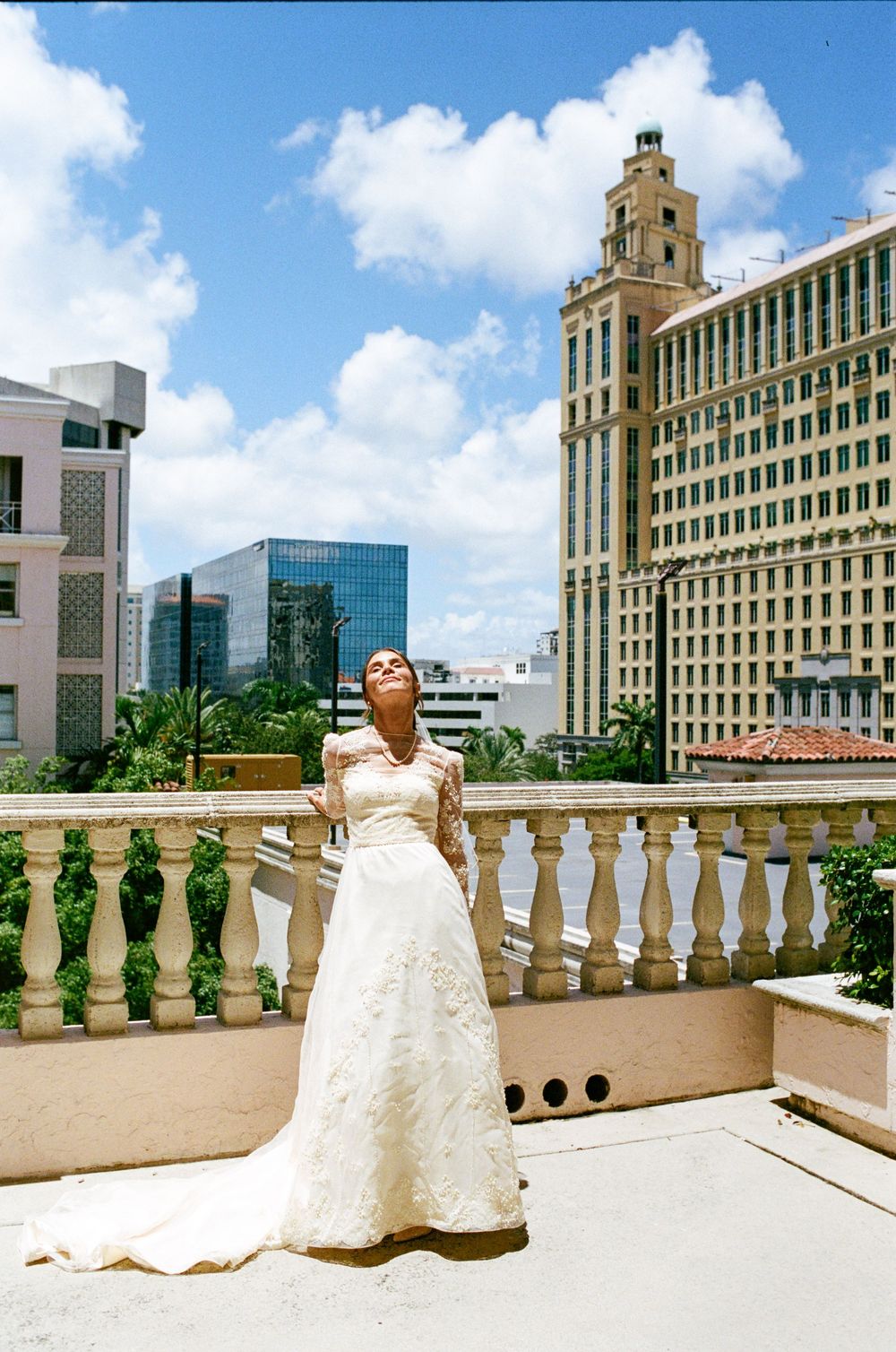 Bride in elegant white lace wedding gown poses on rooftop terrace with city skyline backdrop on sunny day.