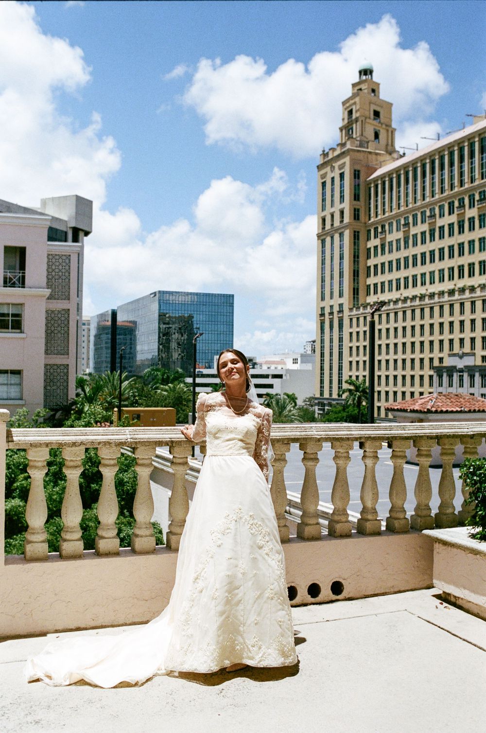Bride in white lace gown poses on rooftop balustrade with city skyline and blue sky backdrop.