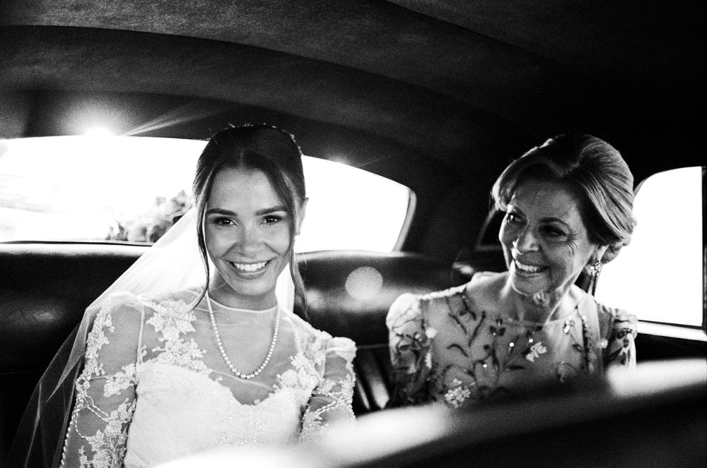Black and white photo of a smiling bride in veil and pearls sitting in a car with an older woman.