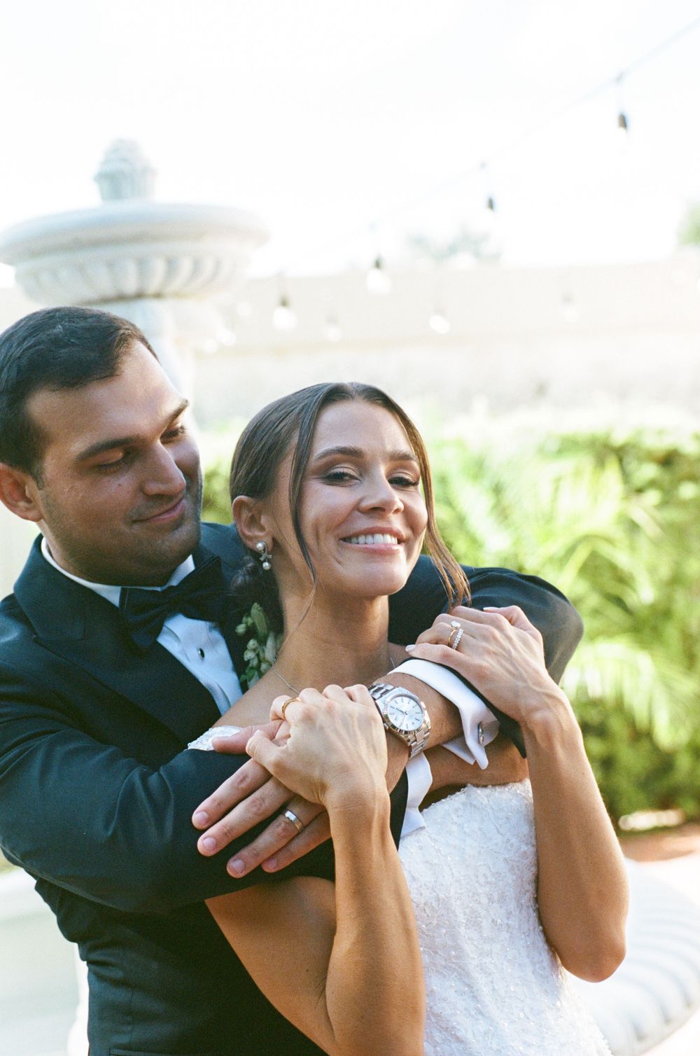 Groom in black tuxedo embraces smiling bride in white dress near outdoor fountain at wedding reception.