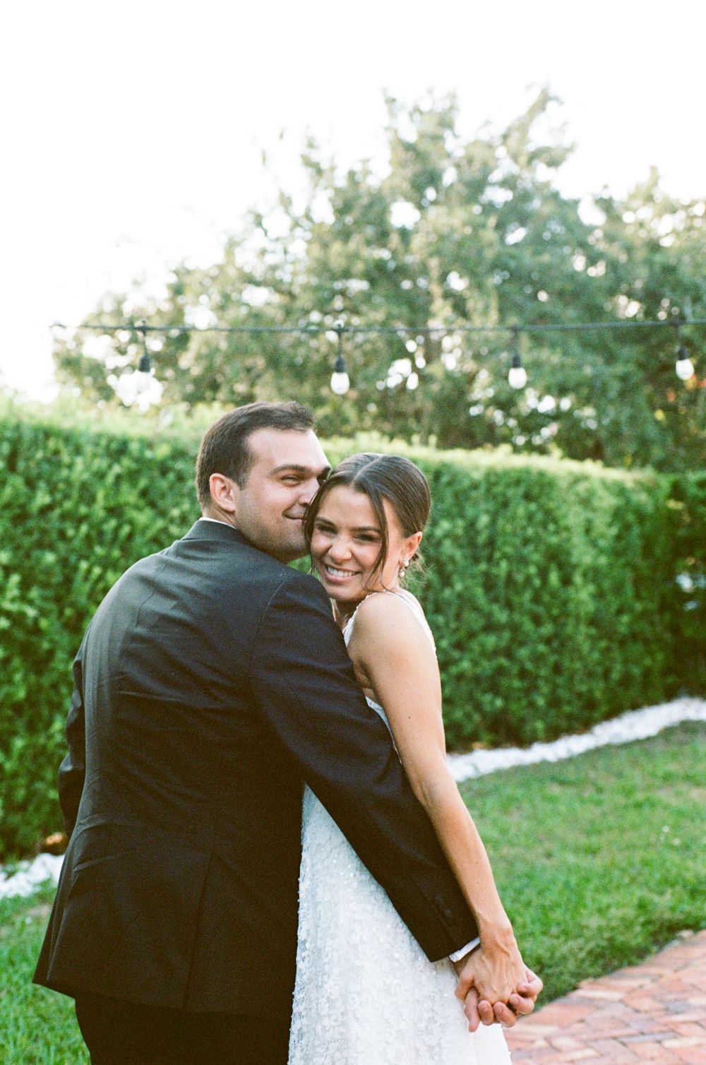 A happy couple embraces outdoors on their wedding day, the groom in a black suit hugging the smiling bride in a white gown.