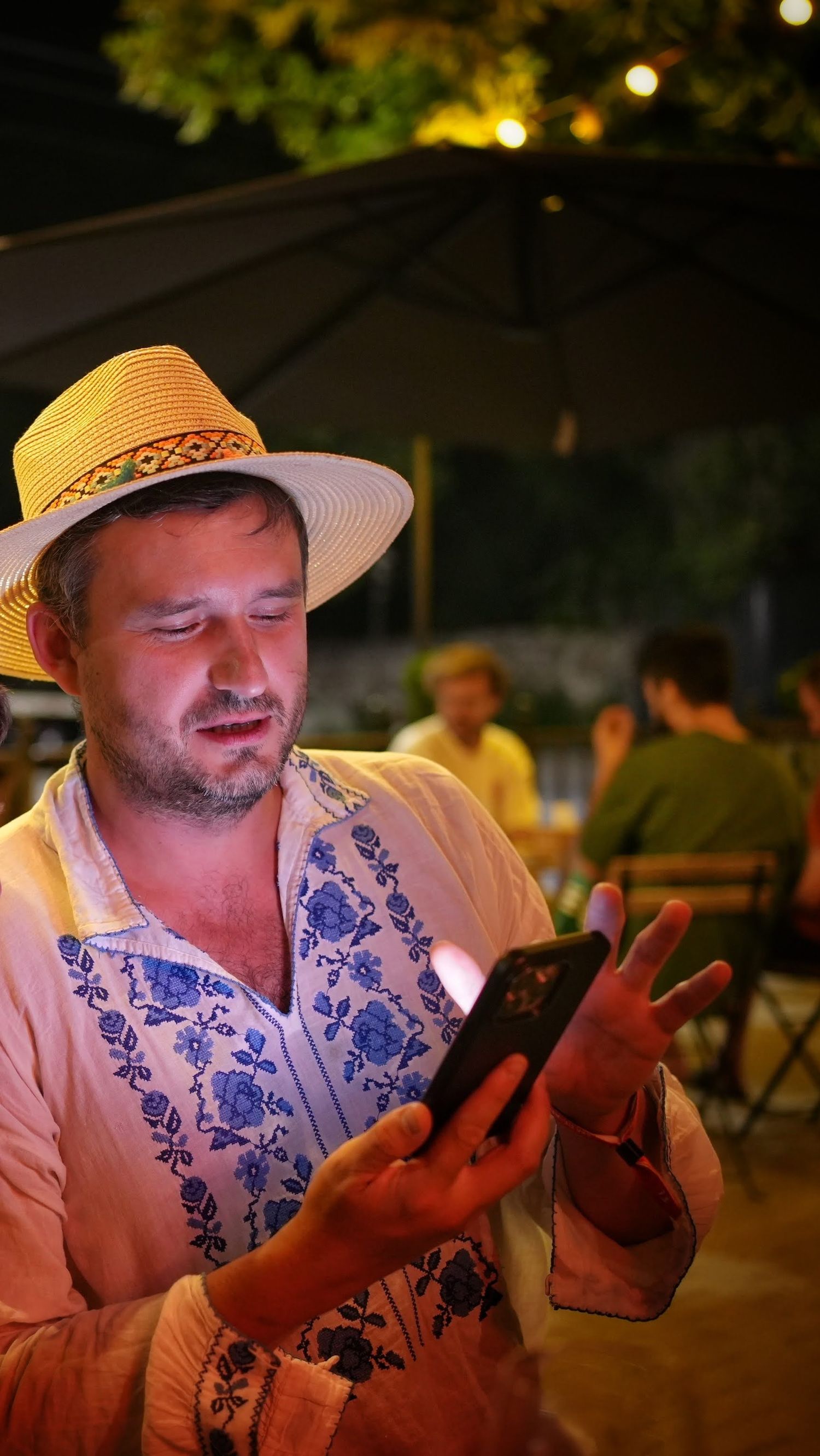 Man wearing a straw hat and embroidered shirt using a smartphone at an outdoor evening venue.
