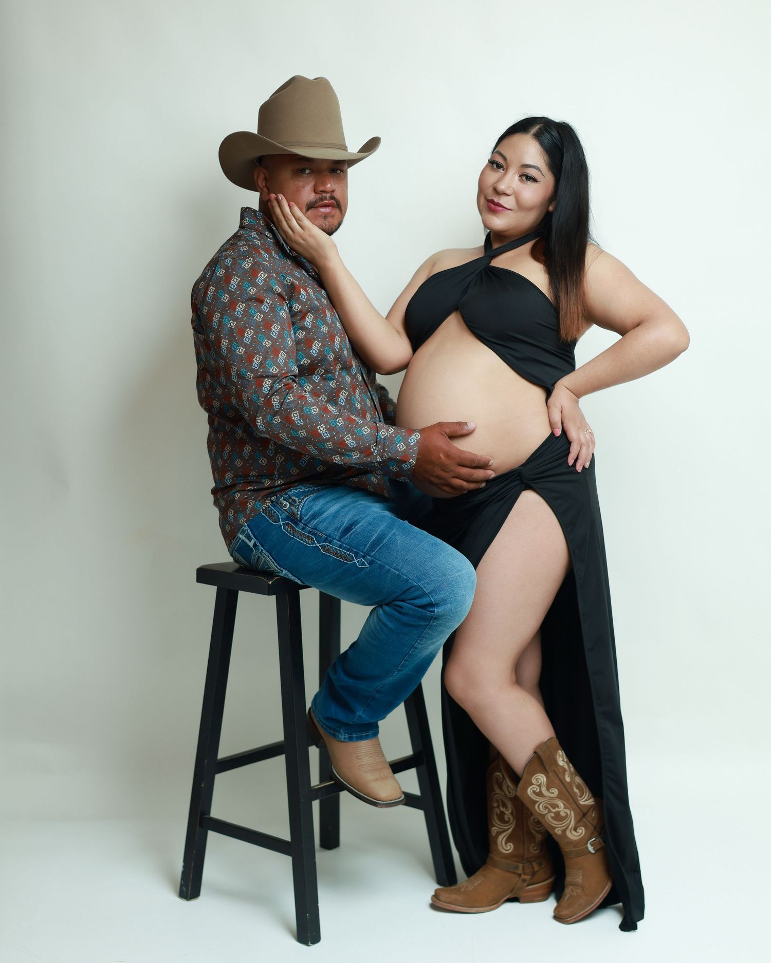 Couple maternity photo with man in cowboy hat and boots sitting on stool beside pregnant woman in black dress.