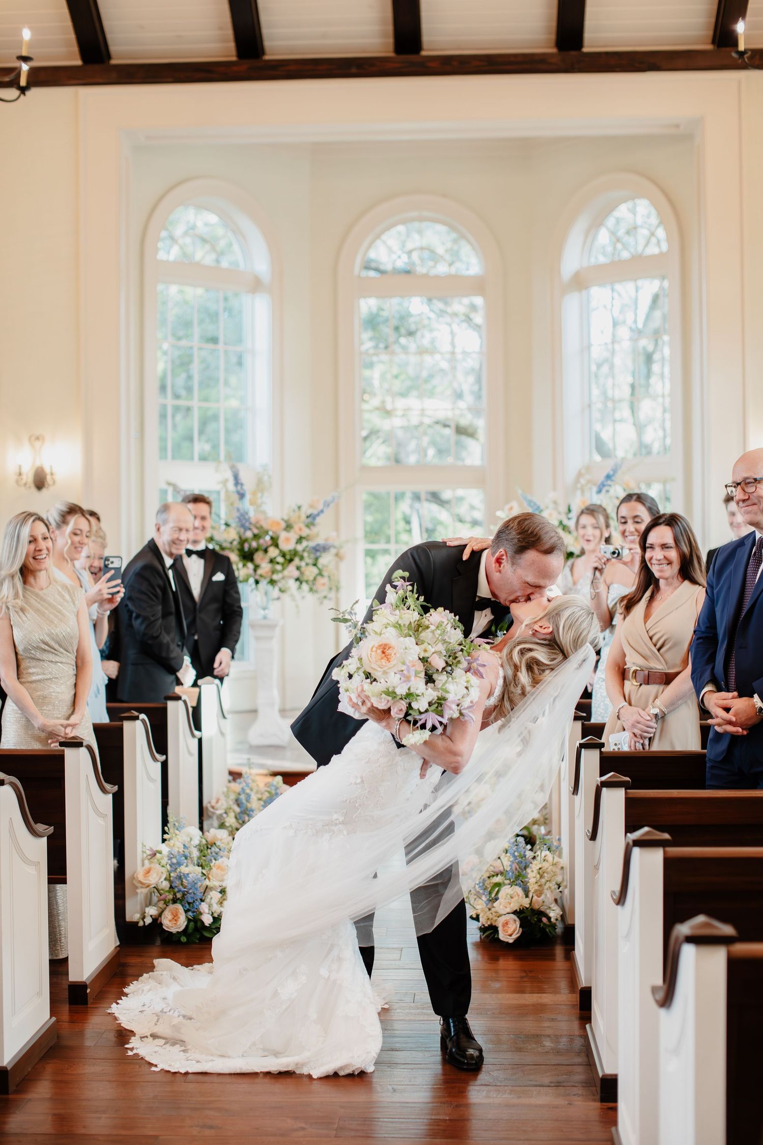 Bride and groom share a romantic dip kiss at the altar in an elegant chapel with floral arrangements and smiling guests.