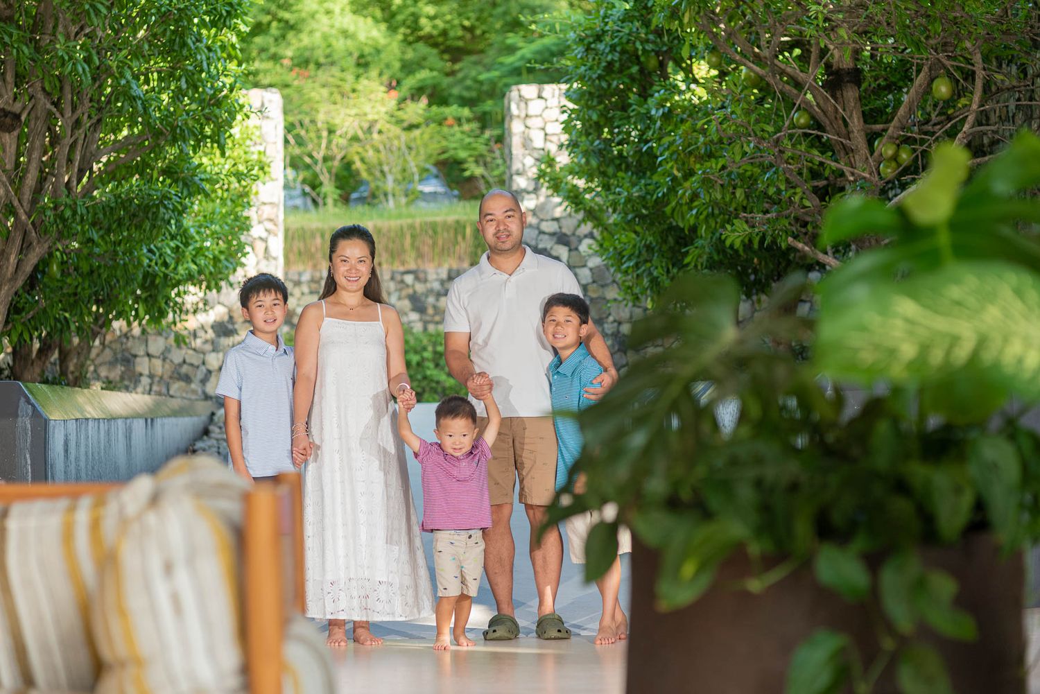 A happy family of five poses together outdoors surrounded by lush green tropical trees and plants.