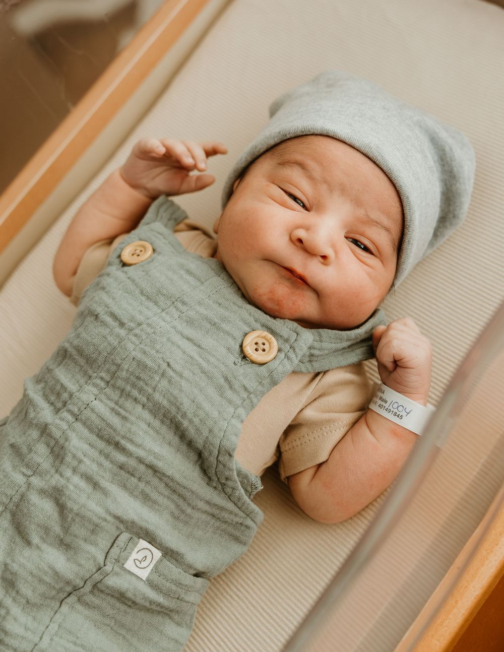 Newborn baby smiling in a hospital bassinet, wearing a sage green linen romper and gray knit beanie hat.