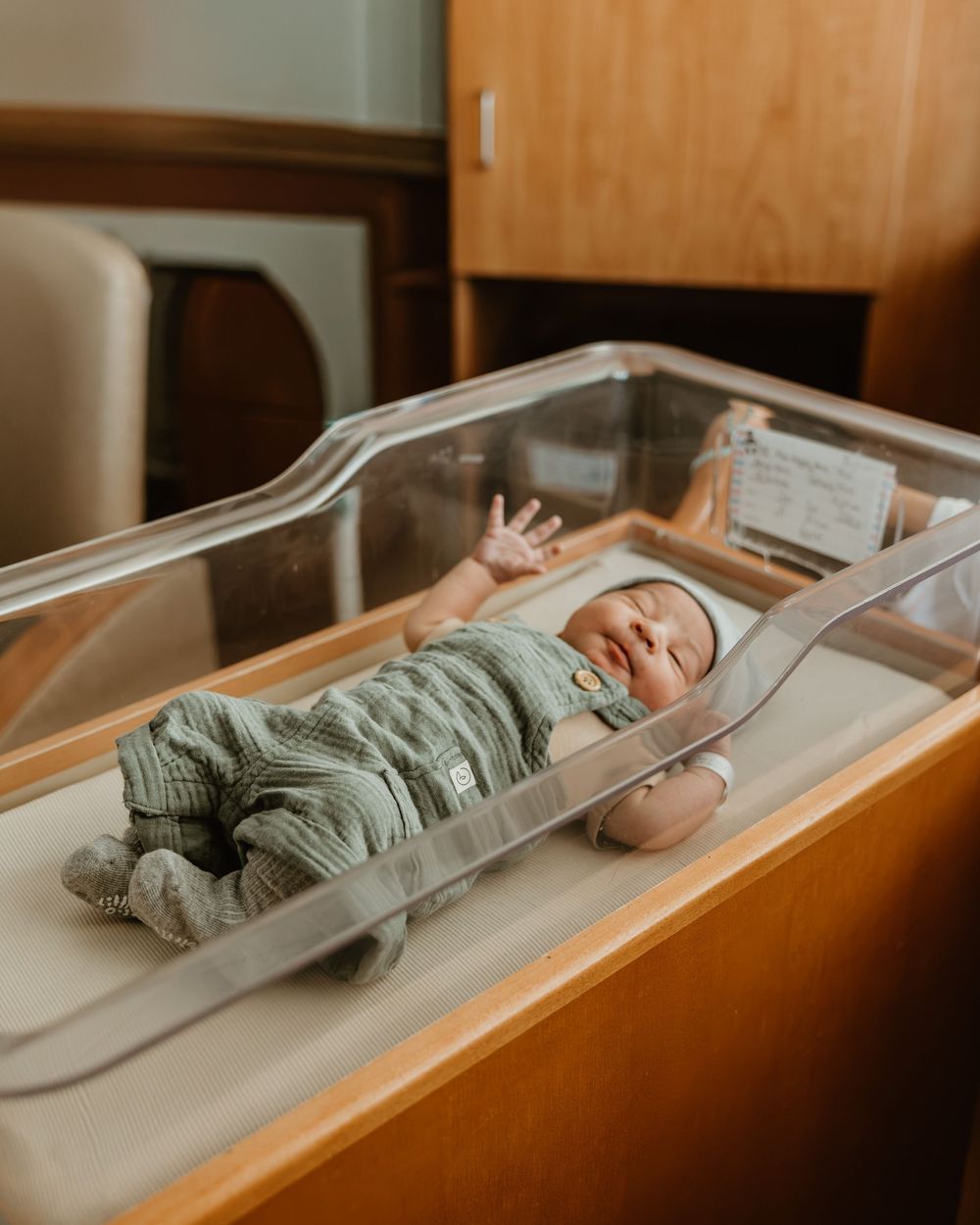 Newborn baby in green knit outfit lying in clear hospital bassinet, raising tiny hand, in a warm wooden room.