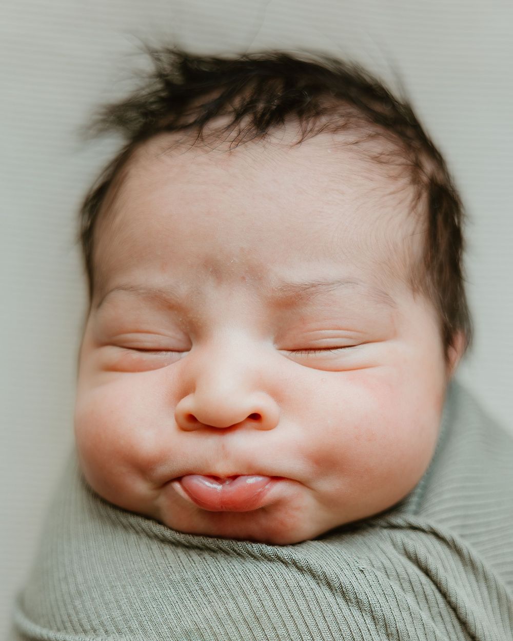 Adorable sleeping newborn baby with dark hair, chubby cheeks, and pouty lips, swaddled in a soft green wrap.