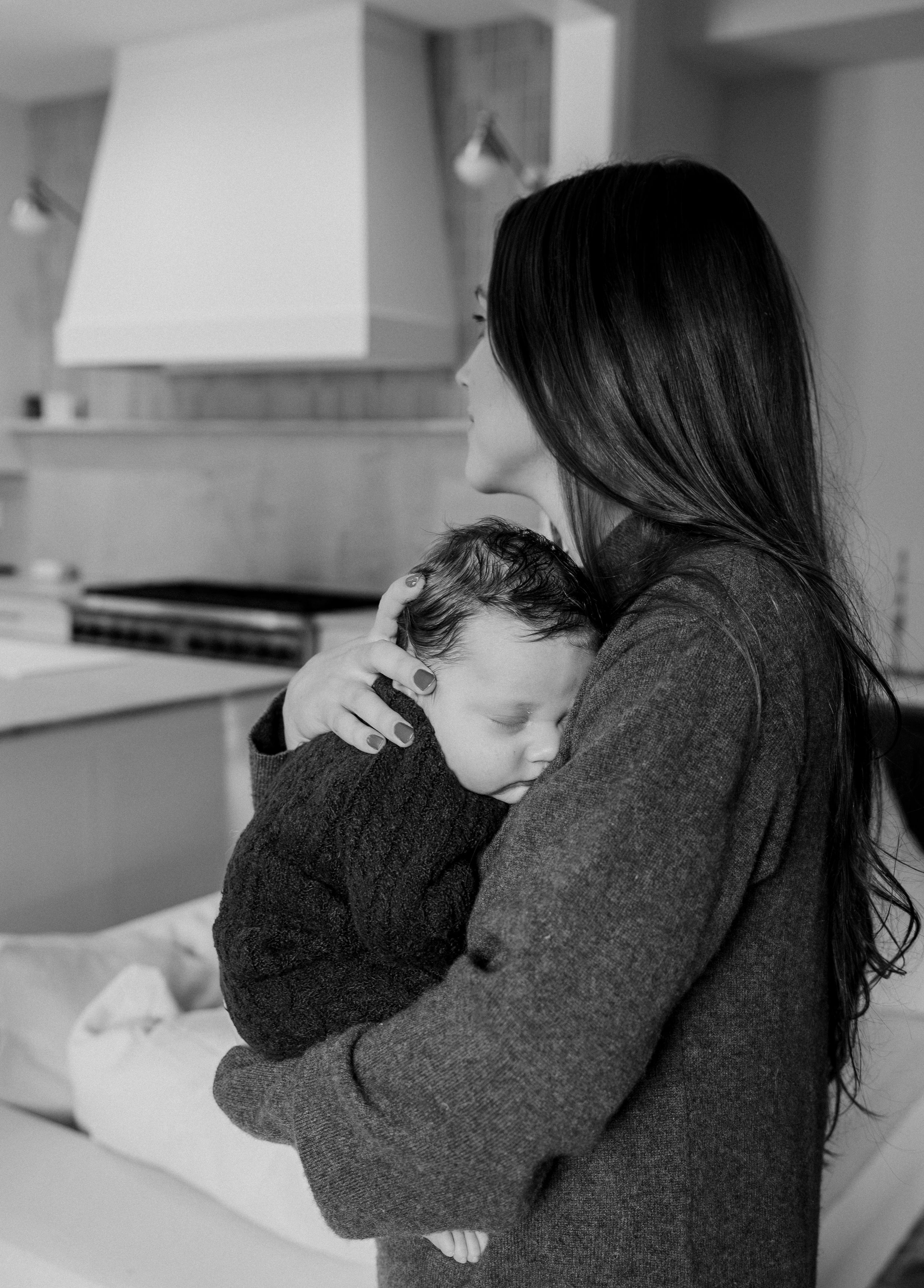 Black and white photo of a mother cradling her sleeping newborn baby in a modern kitchen.