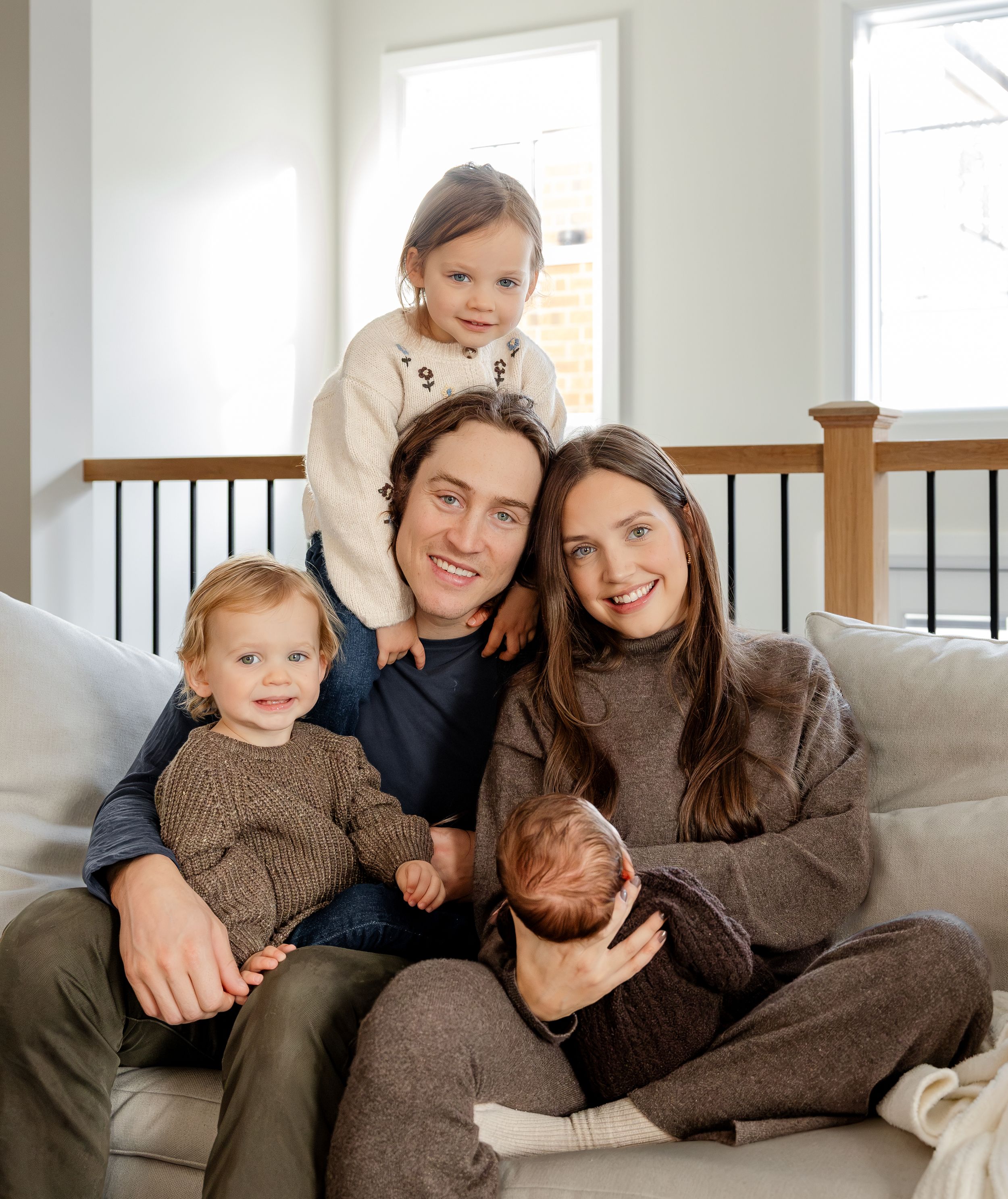 Happy family of five posing on a couch, including two toddlers and a newborn baby, in a bright modern home.