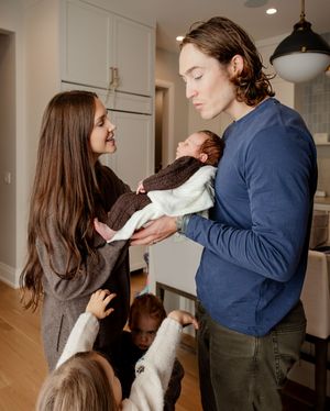 A man in a blue shirt and woman with long dark hair admire a newborn baby indoors while children gather around them.