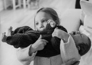 Black and white photo of a toddler holding a newborn baby wrapped in a dark blanket with a pacifier.