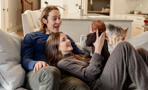 Family relaxing on couch together laughing with a small dog in a cozy living room.