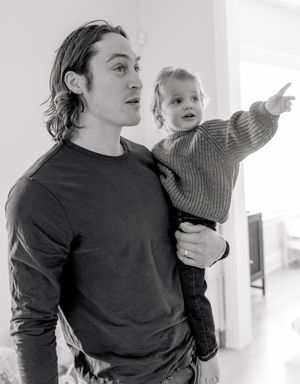 Black and white photo of a young man holding a toddler who points at something off-camera indoors.