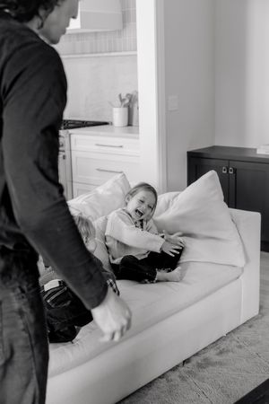 Black and white photo of a laughing toddler on a couch with an adult standing nearby in a cozy home.