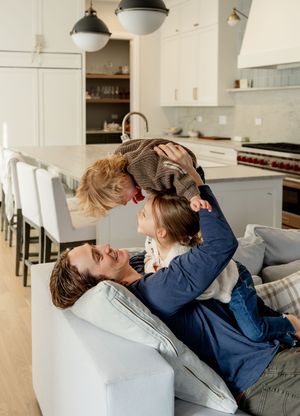 A father lies on a couch lifting his young child while another child sits nearby in a bright modern kitchen home.
