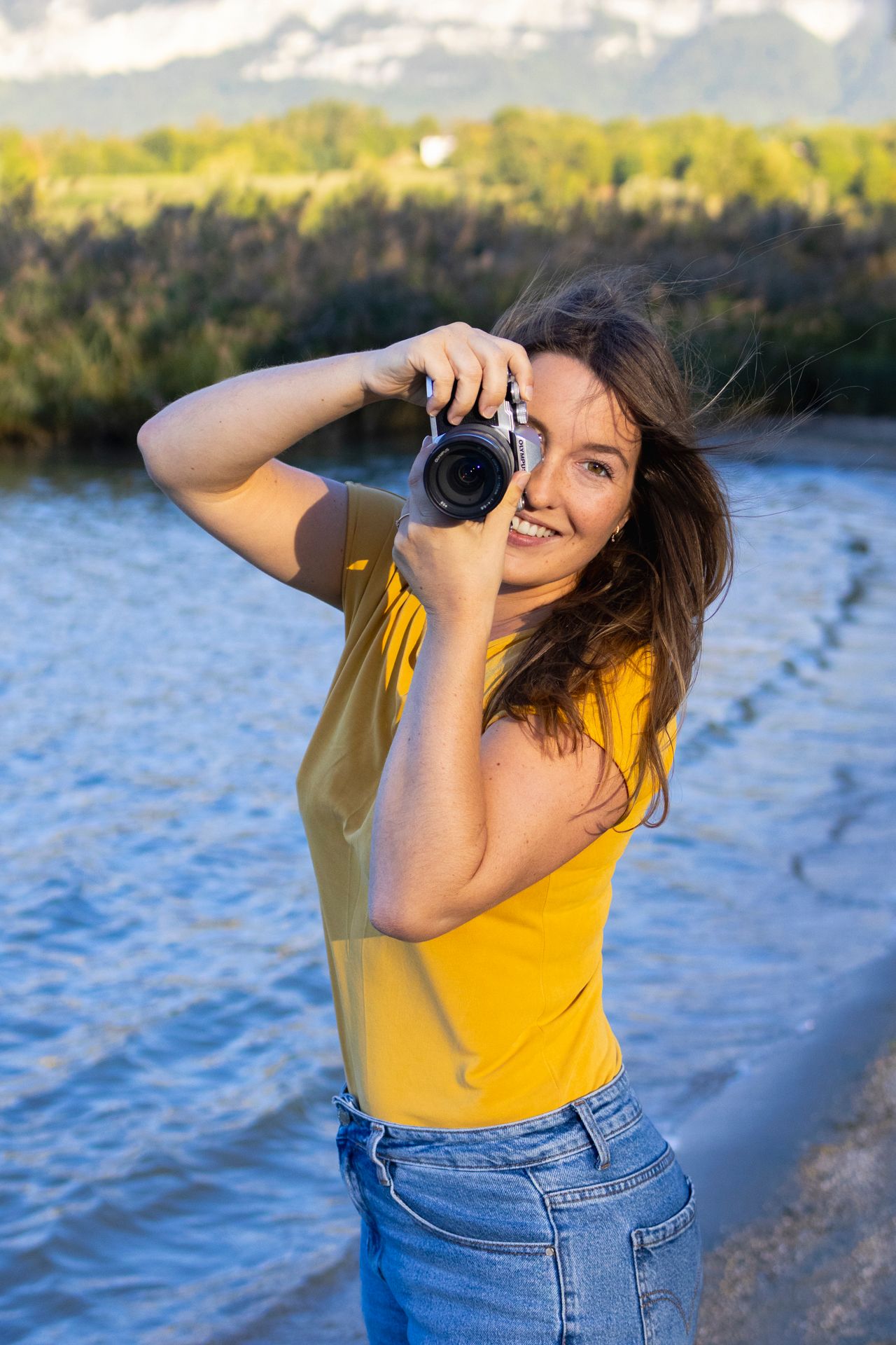 Jade, photographe, en haut jaune, appareil photo en main au bord de l’eau, lors d’une séance en Savoie