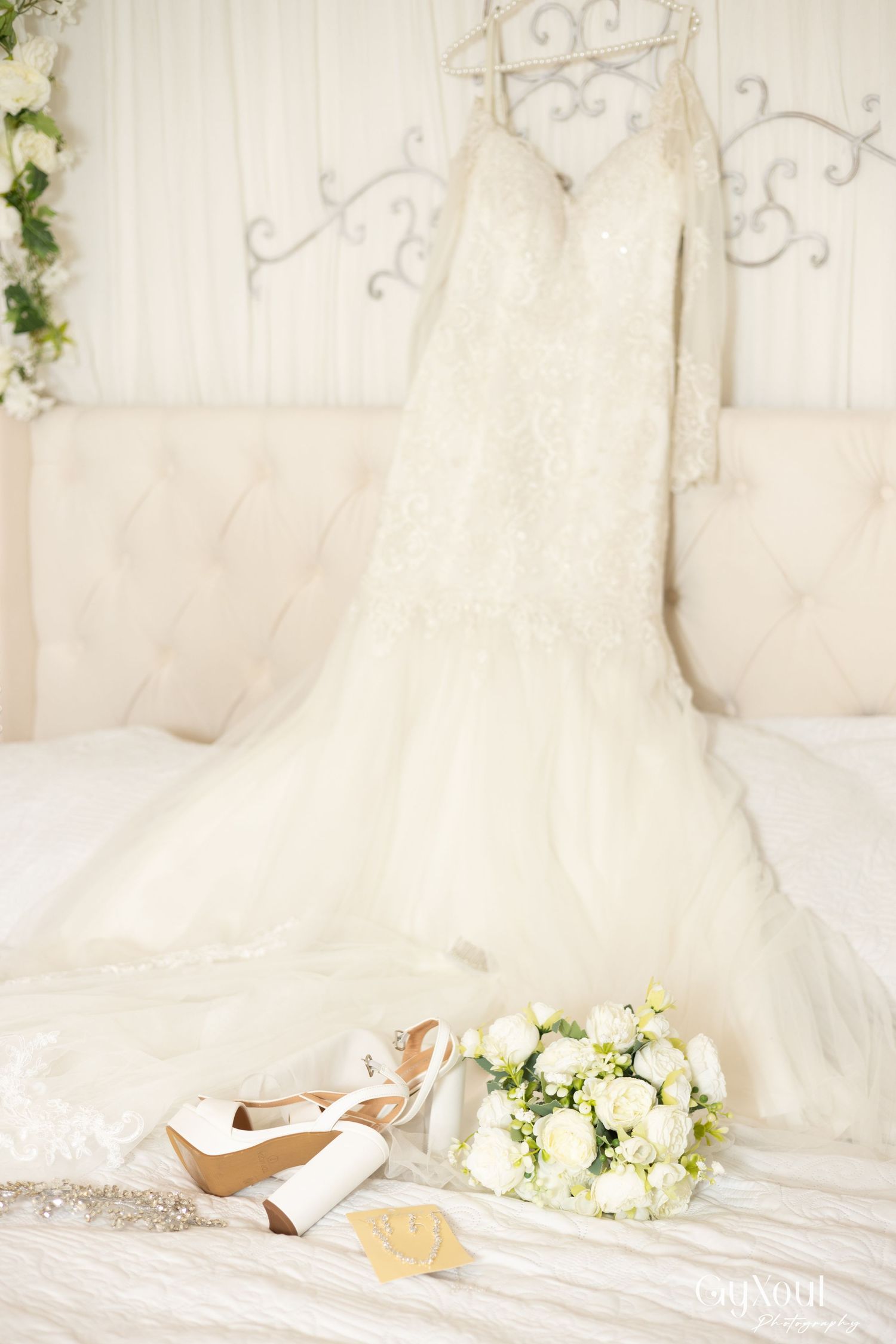 Elegant mermaid wedding dress hanging on ornate headboard with white bridal heels and rose bouquet on bed.