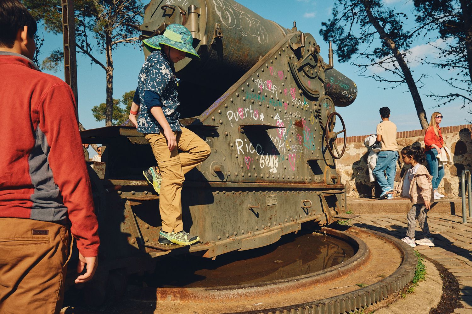 People climbing on a graffiti-covered military cannon exhibit outdoors on a sunny day.