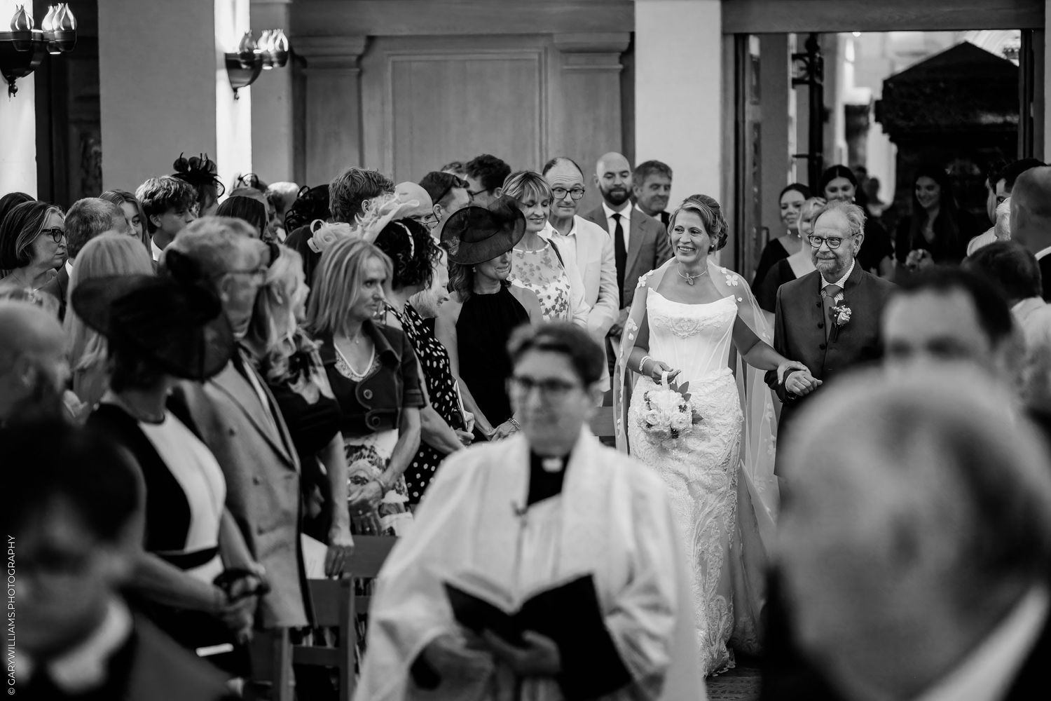 Bride walks down the aisle with escort at elegant church wedding ceremony, guests seated in pews, black and white photo.