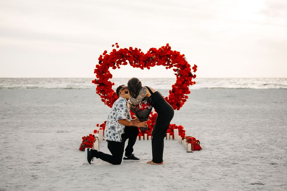 Romantic beach proposal with red rose heart arch, candles, and couple embracing on sandy shore at sunset.