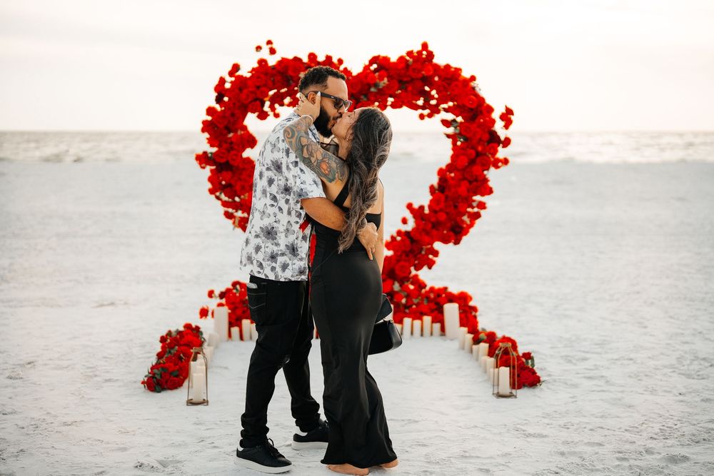 Couple kissing in front of red rose heart arch on beach with candles at romantic proposal setup.