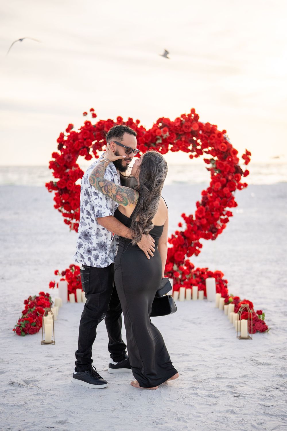 Couple kissing in front of a large red rose heart arch with candles on a beach at sunset.