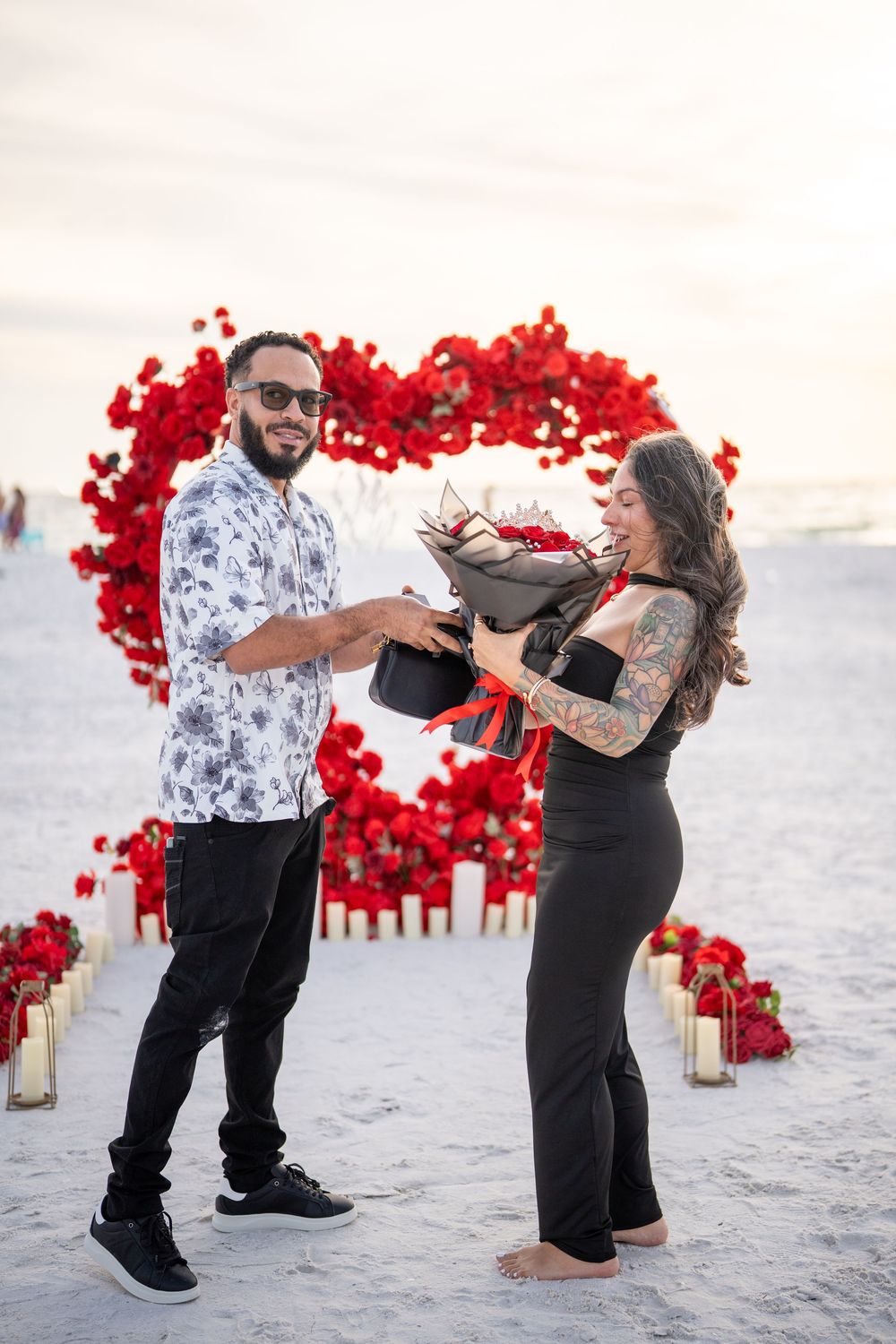 Couple exchanging red rose bouquet on beach with heart-shaped floral arrangement and candles at sunset.
