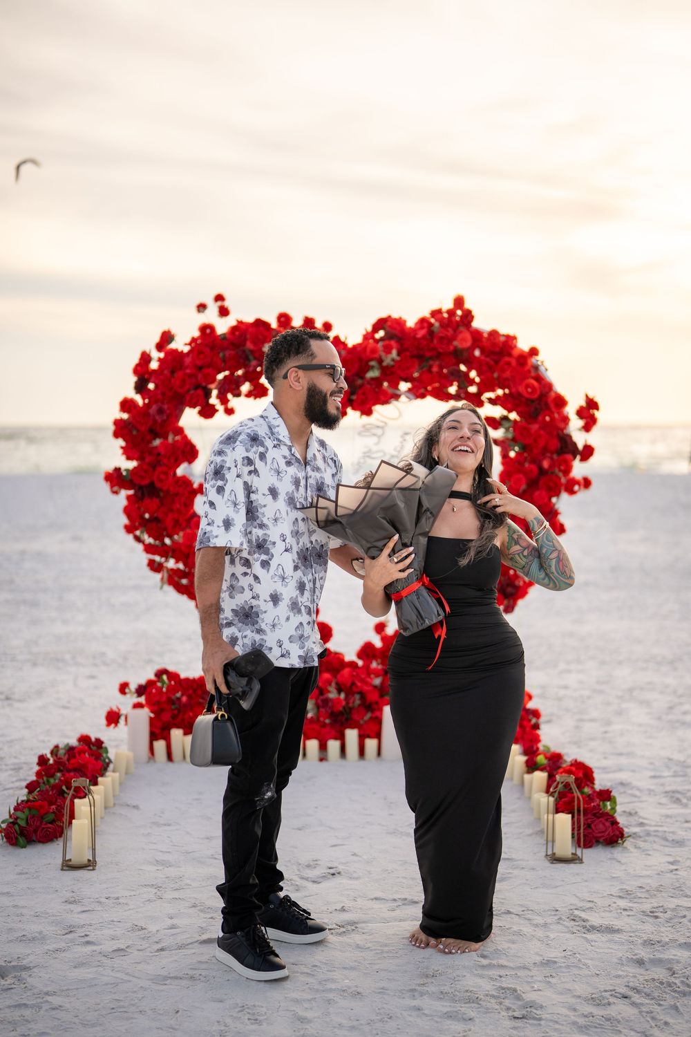 Couple laughing on beach with red rose heart arch, candles, and bouquet at romantic sunset proposal setup.