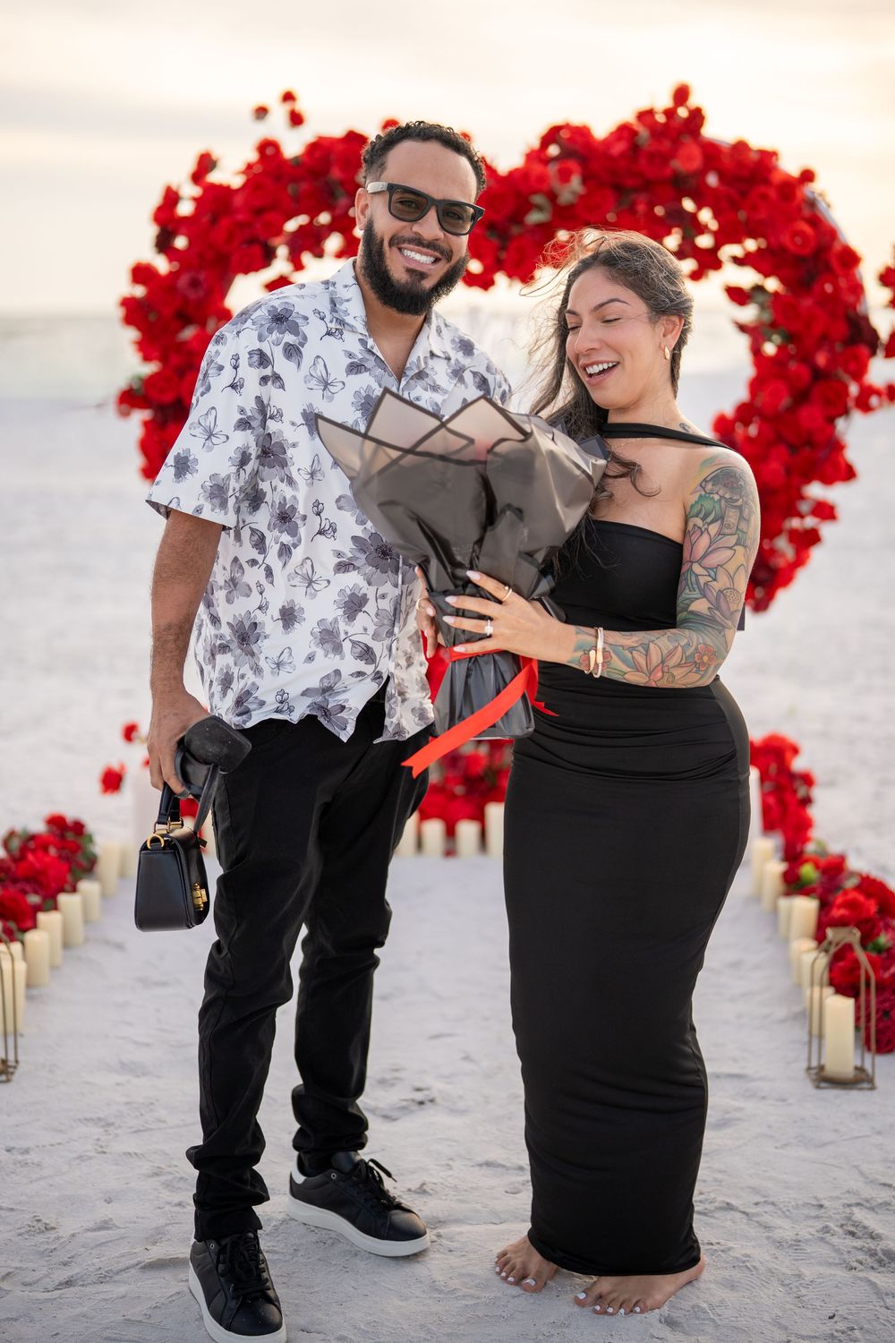 Couple smiling at beach proposal setup with red rose heart arch and candles at sunset.