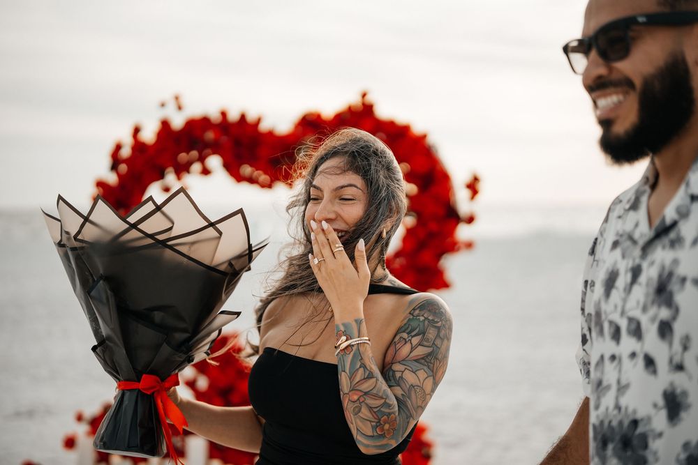Woman with tattoos laughing holding flower bouquet during romantic beach proposal with red heart backdrop.