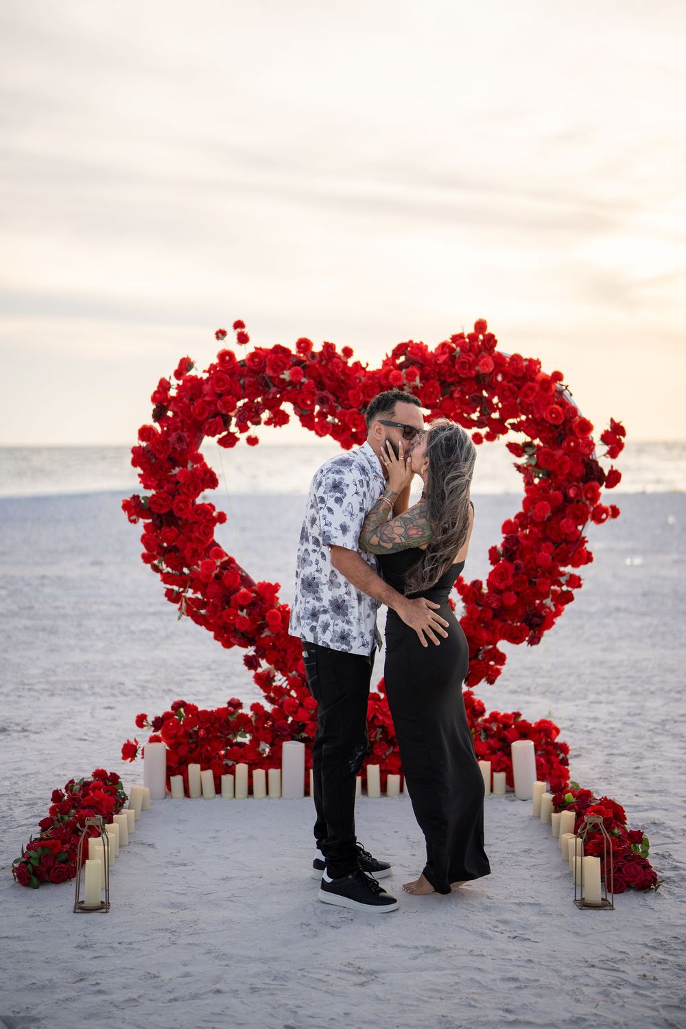 Couple kissing in front of a large red rose heart arch on a beach at sunset with candles lining the path.
