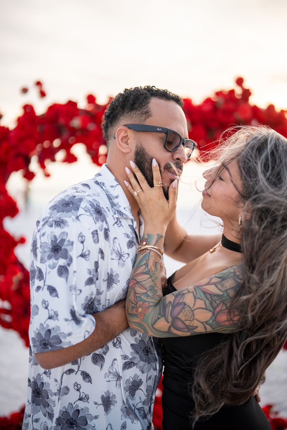 Couple sharing a romantic moment in front of a red floral heart backdrop, woman with tattoos and gray hair touching mans face.