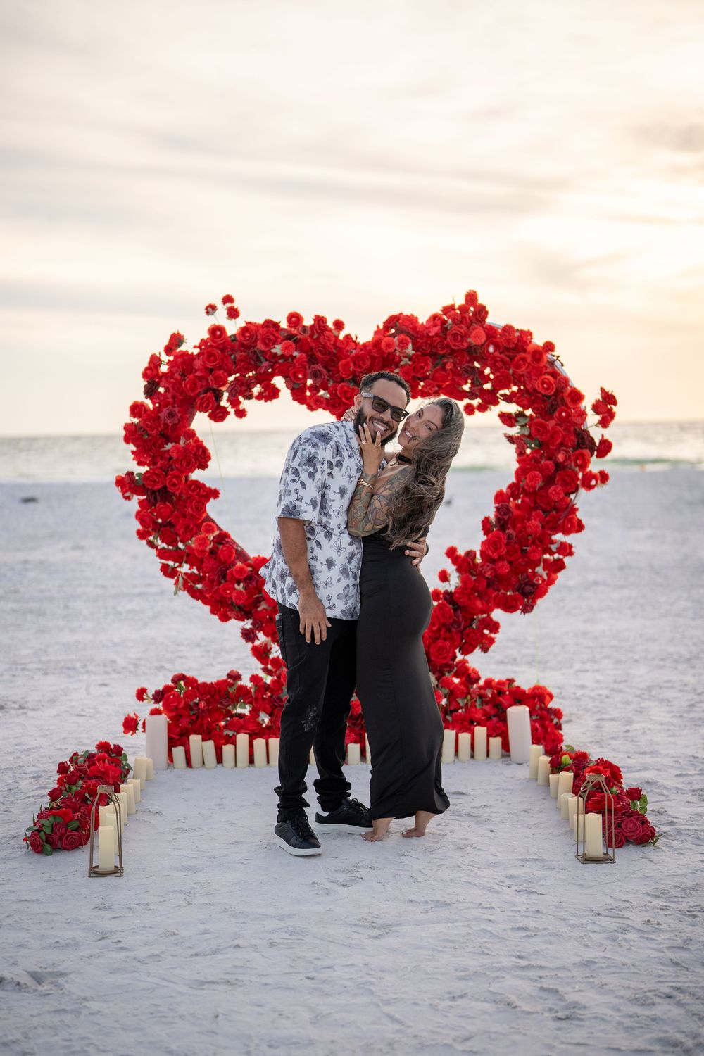 Couple embracing in front of a large red rose heart sculpture on a beach at sunset with candles.