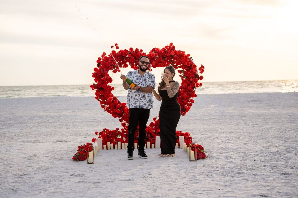 Couple celebrating beach proposal with red rose heart arch, candles on sand at sunset.
