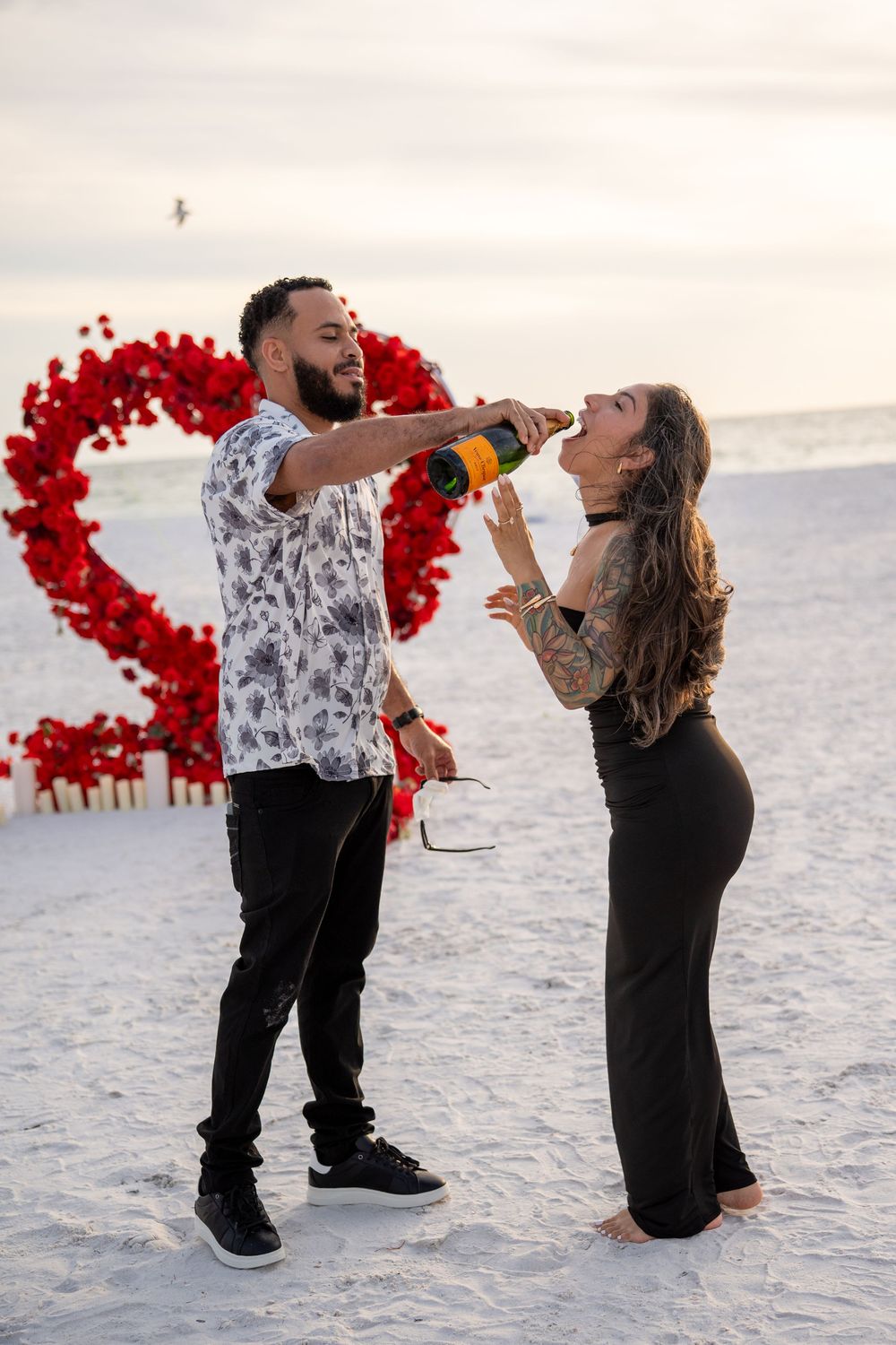 Couple celebrating on a beach with champagne, red heart floral backdrop, romantic Valentine's Day proposal setup at sunset.
