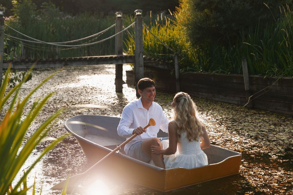 Couple sharing a romantic boat ride on a sunlit pond surrounded by lush greenery and a wooden bridge.