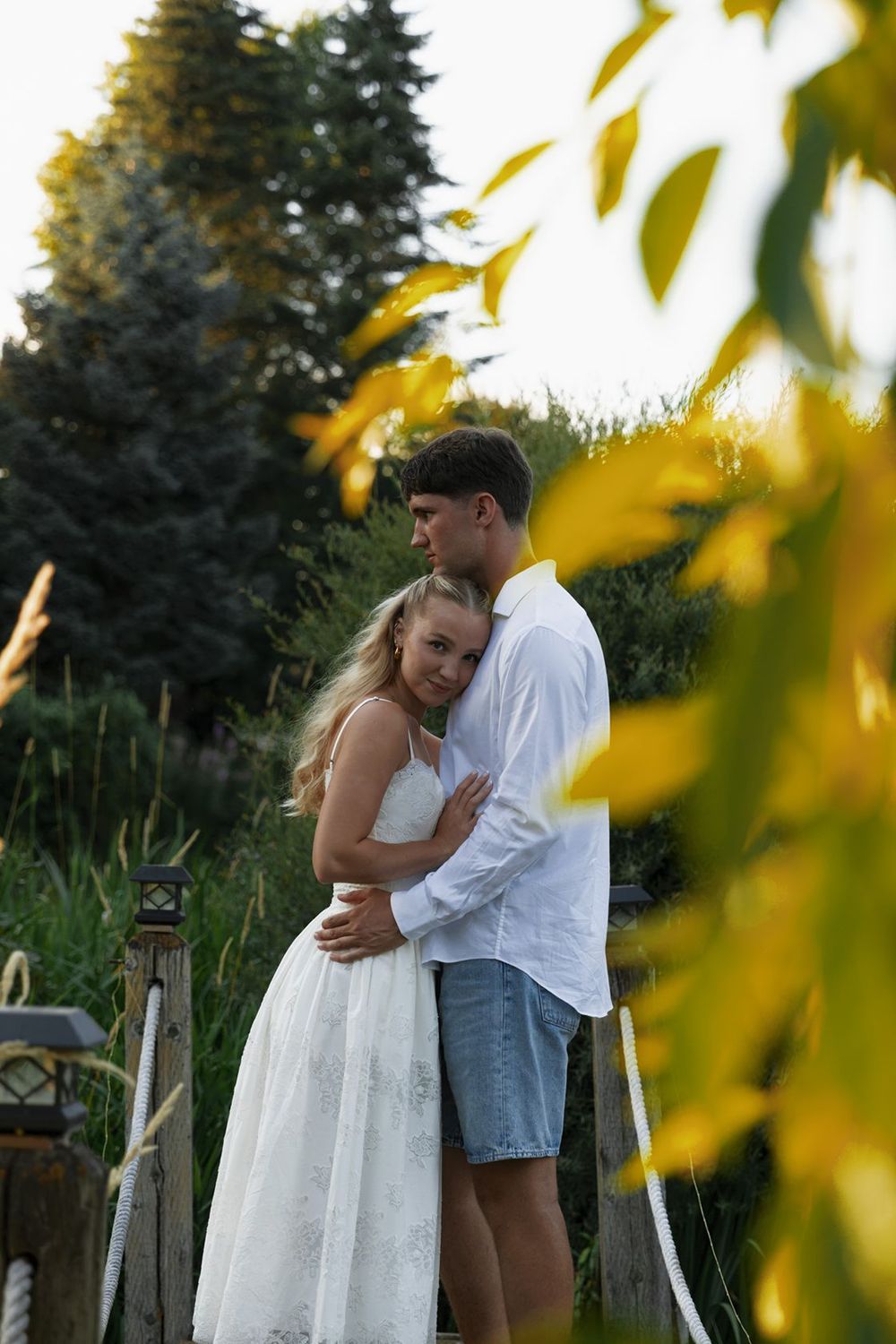 Couple embracing outdoors near wooden fence, woman in white dress, man in white shirt and denim shorts, lush green trees background.
