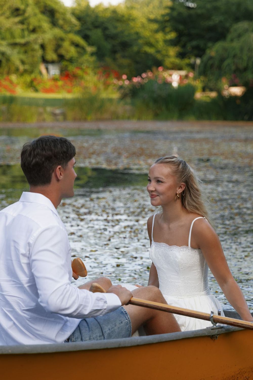A young couple shares a romantic moment in an orange rowboat on a serene lily pad-covered lake.