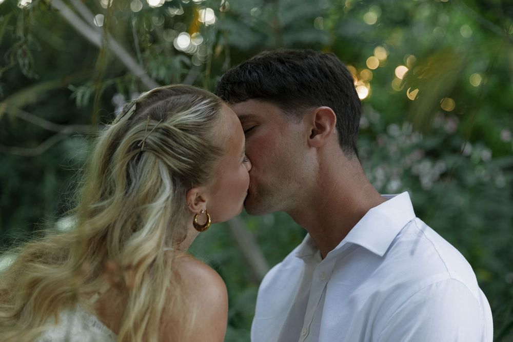 A couple shares a romantic kiss outdoors surrounded by lush green foliage.
