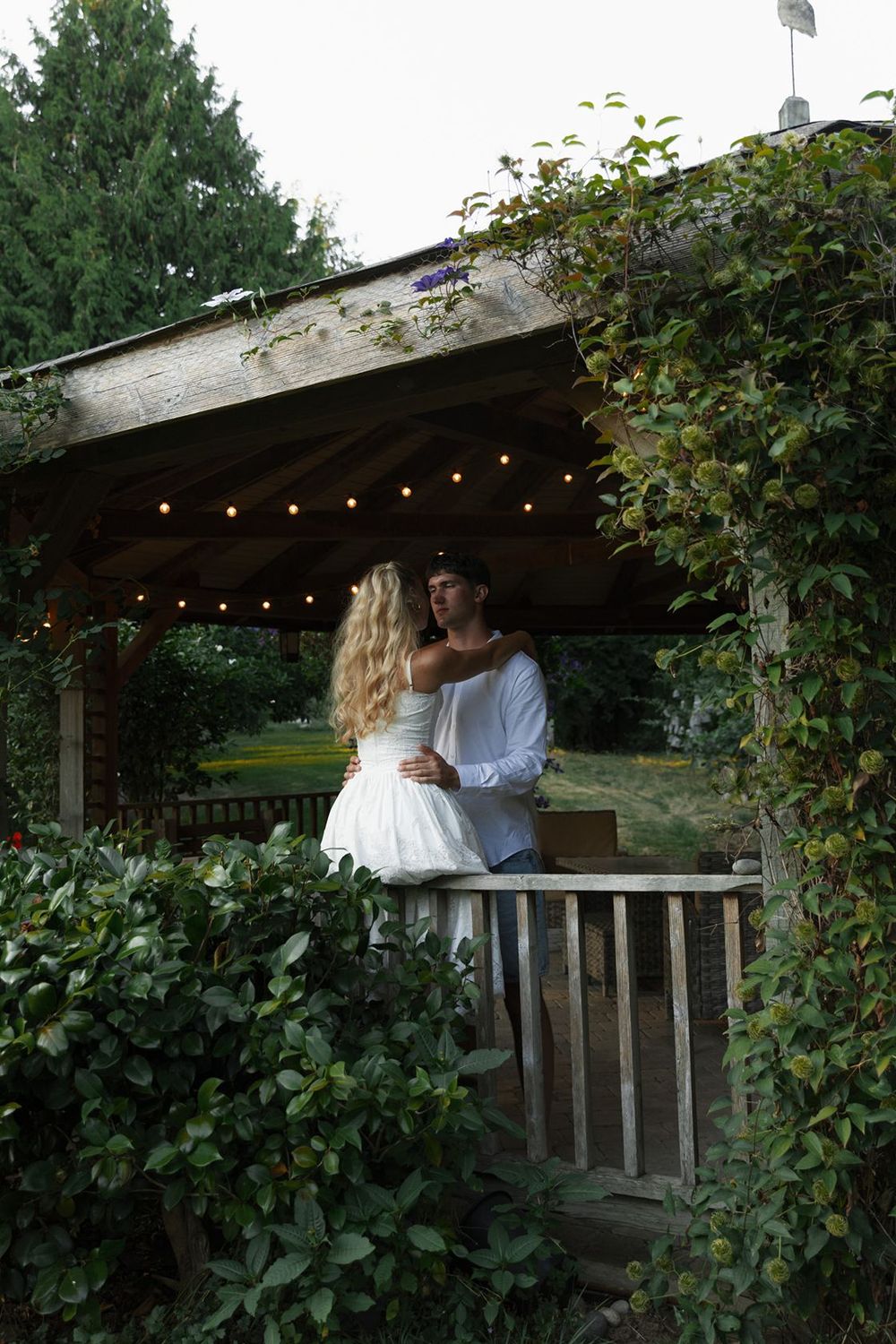 Couple embracing romantically under a rustic garden gazebo with string lights, surrounded by lush greenery at dusk.