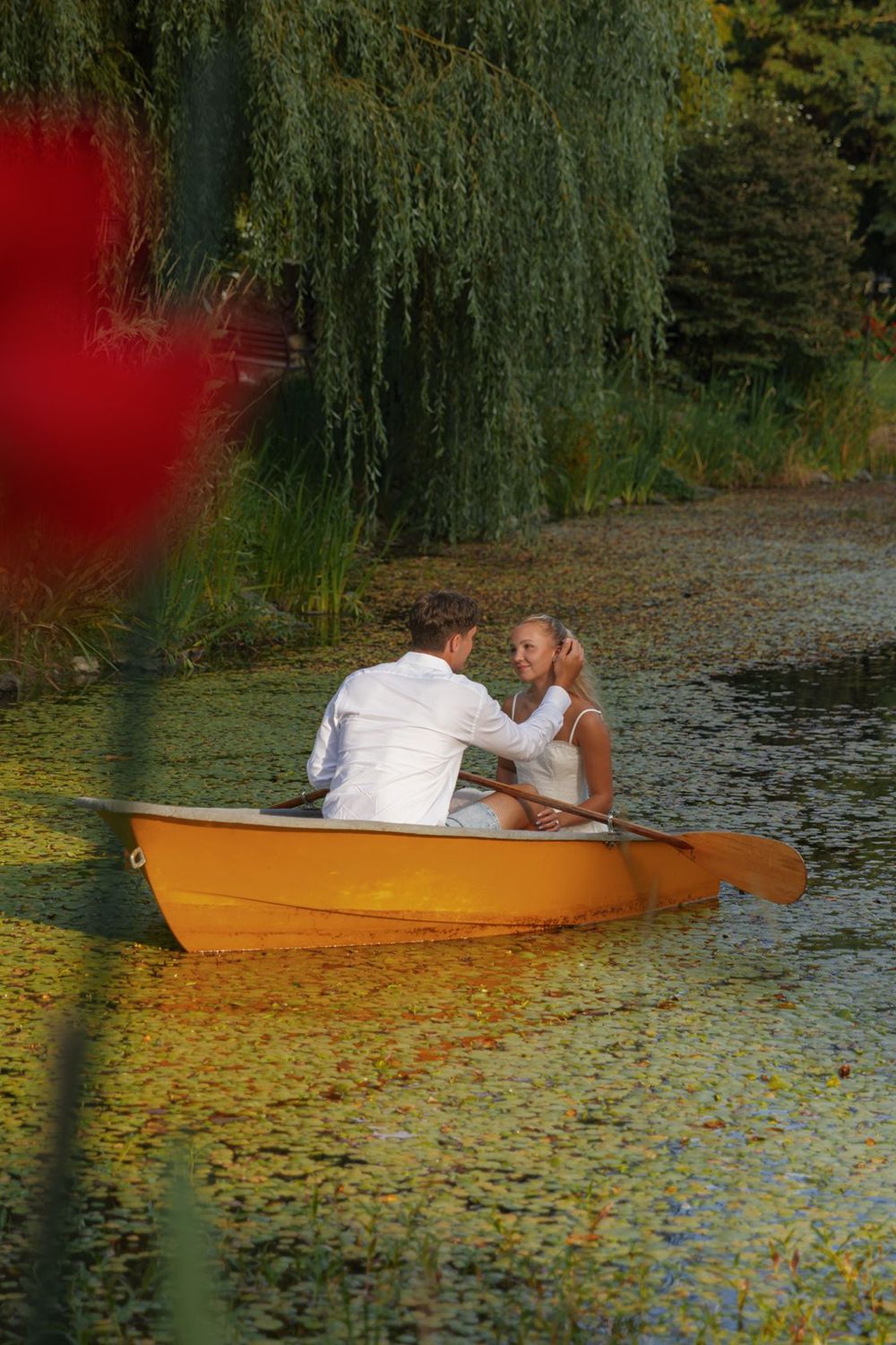 A couple enjoys a romantic boat ride in an orange rowboat on a peaceful green lake surrounded by willows.