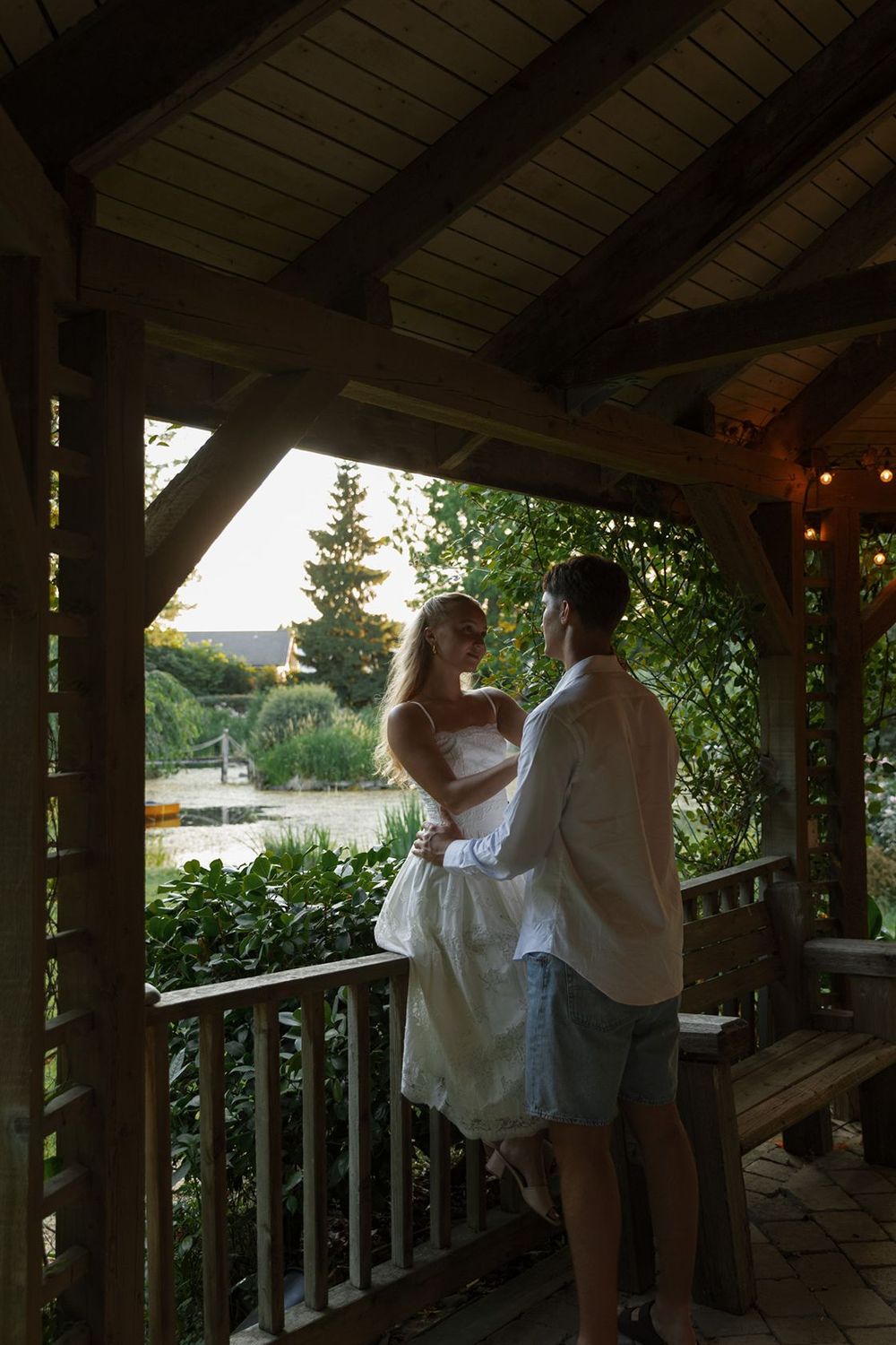 Couple embracing on a wooden gazebo porch surrounded by lush greenery at sunset.