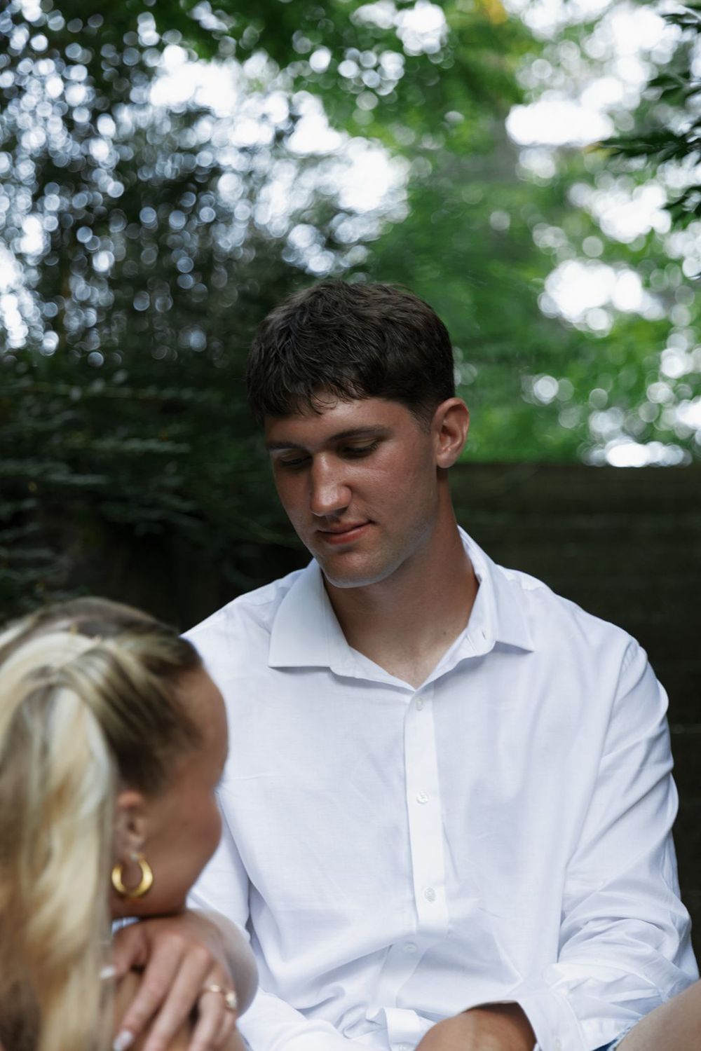 Young man in white shirt looking down outdoors, with blurred green trees in background.