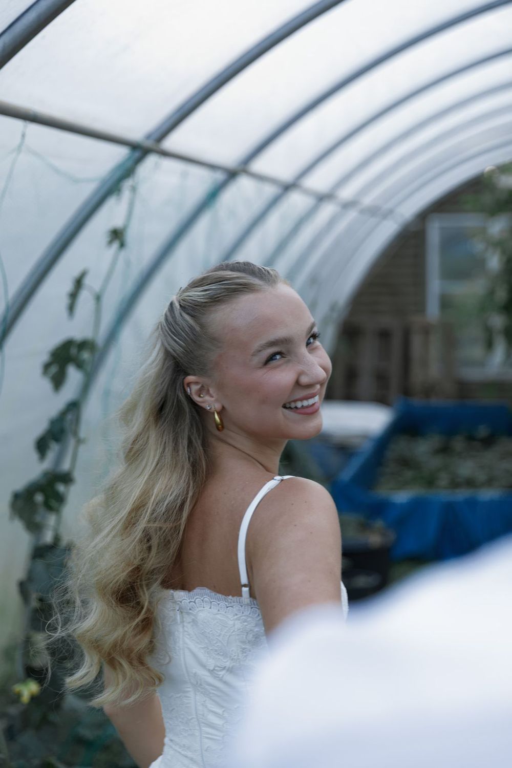 Smiling blonde woman in white dress inside a greenhouse tunnel, looking over her shoulder with long wavy hair.