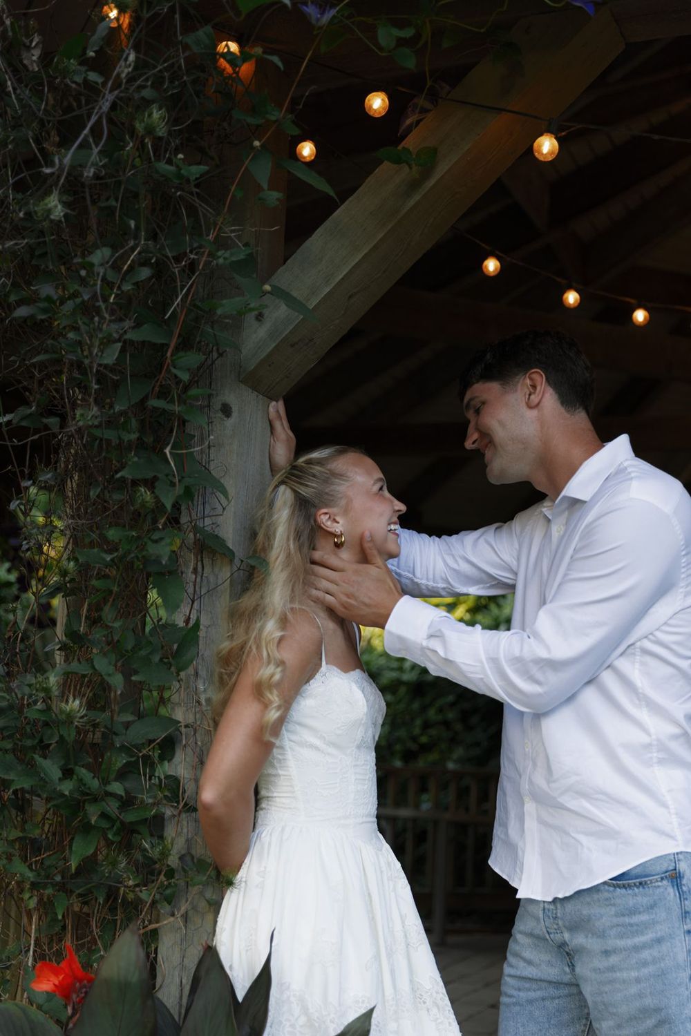 Couple in white outfits sharing a tender moment under string lights on a rustic wooden porch with ivy.