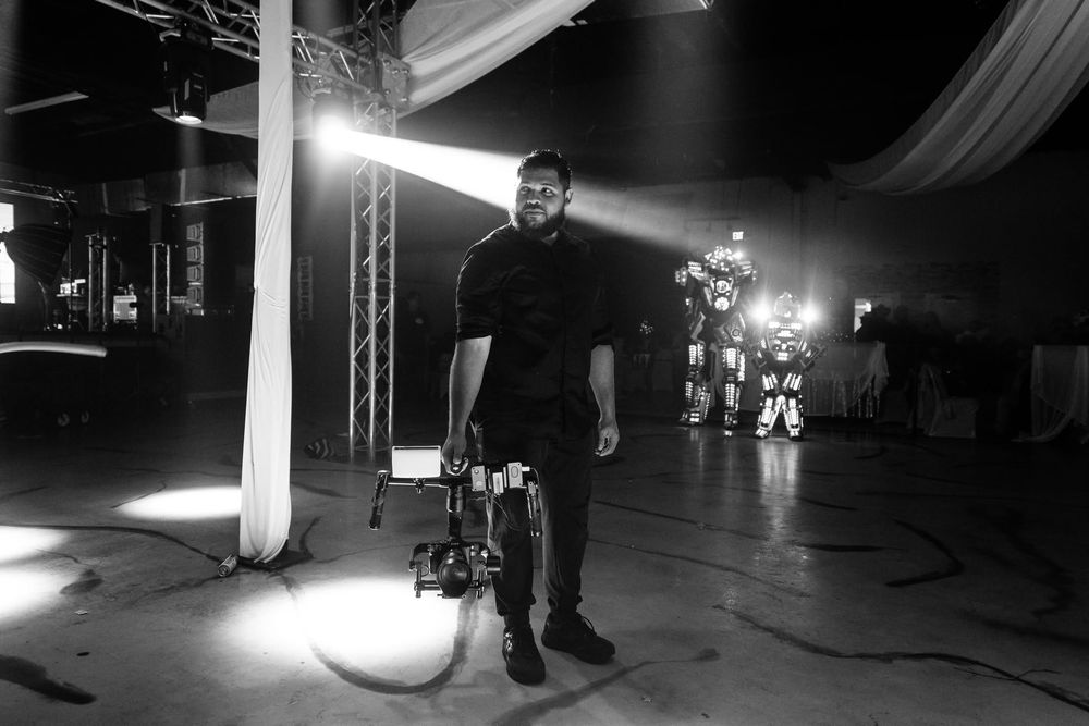 Black and white photo of a man standing near a camera dolly on a film set with bright stage lights in background.