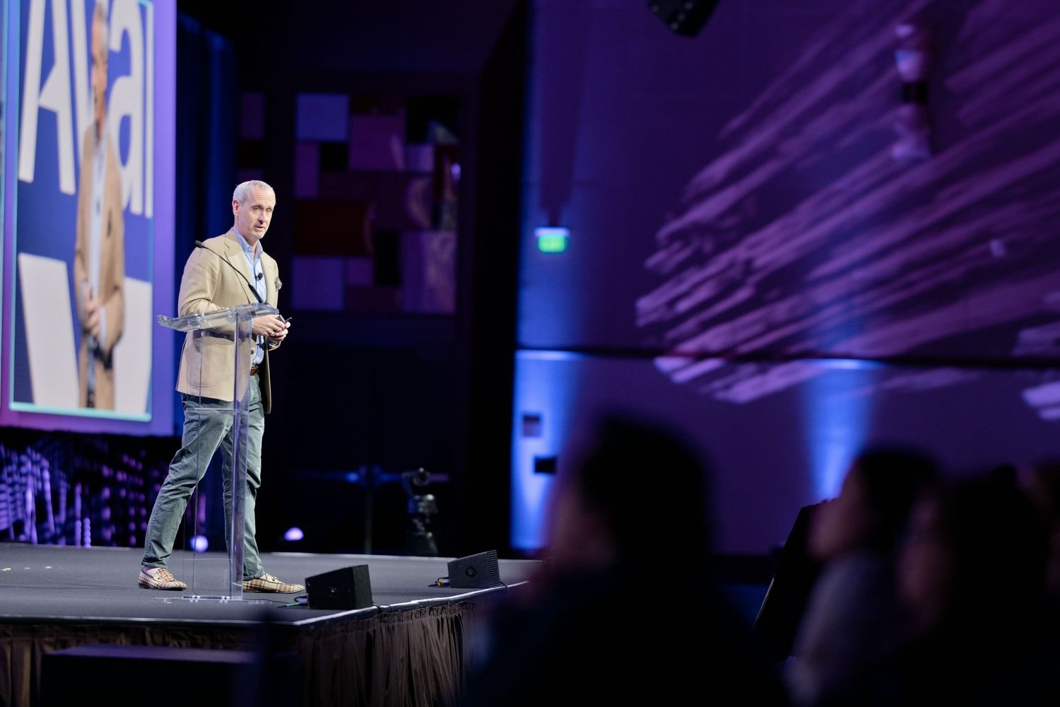 A speaker presents on a purple-lit conference stage to an audience, with a large screen displaying content behind them.